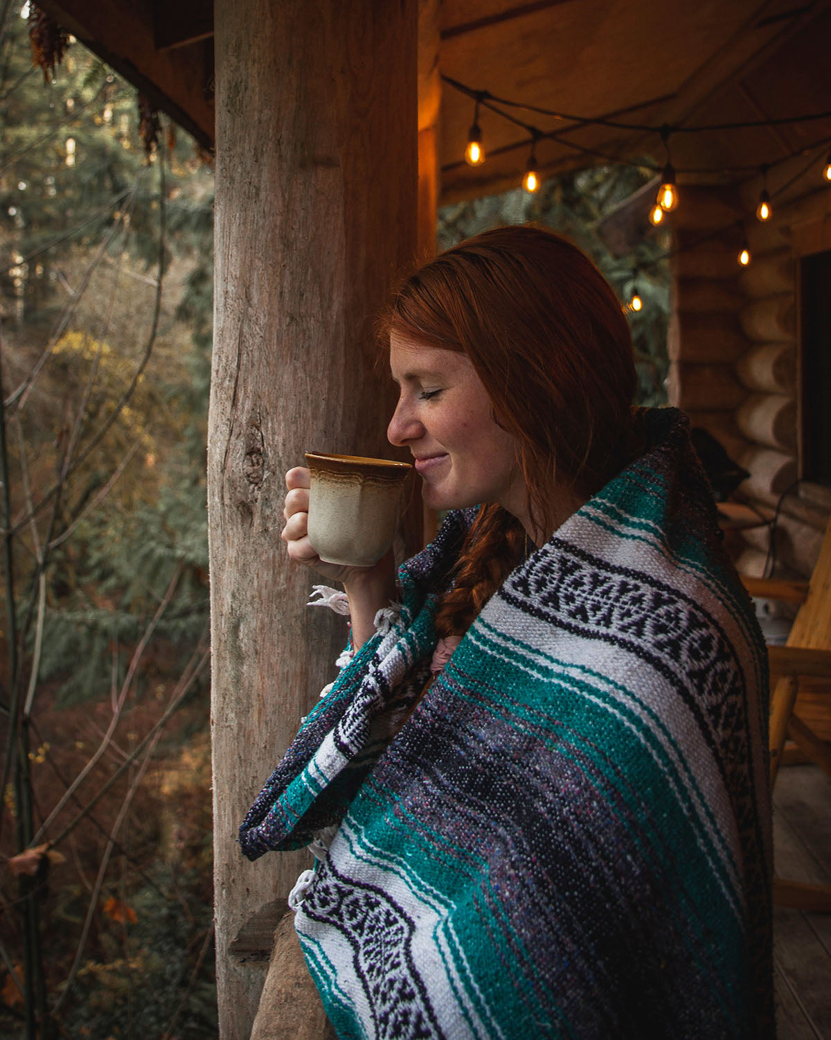 woman with red hair wrapped in blue aztec blanket sipping tea in front of cabin